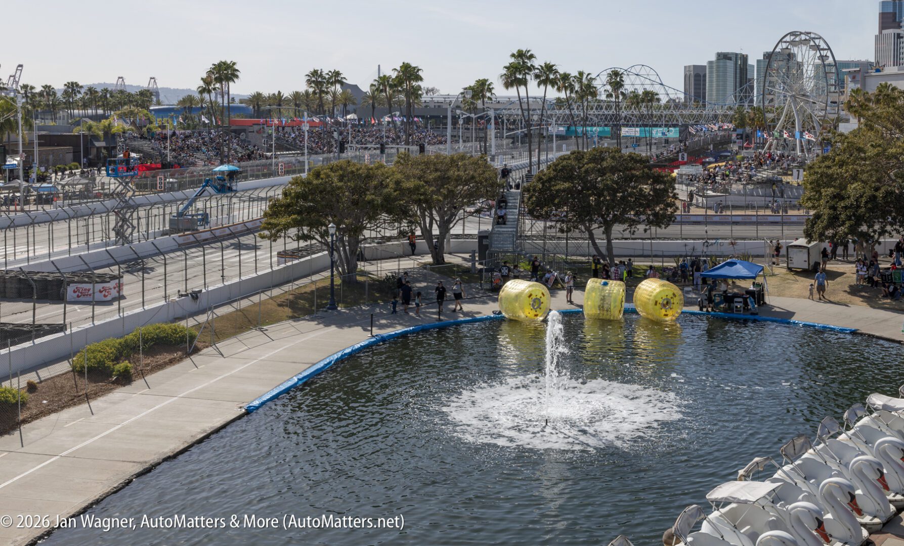 A car race track curves around a pond with a fountain; large yellow inflatable balls float in the water. Spectators sit in stands, and palm trees line the area.
