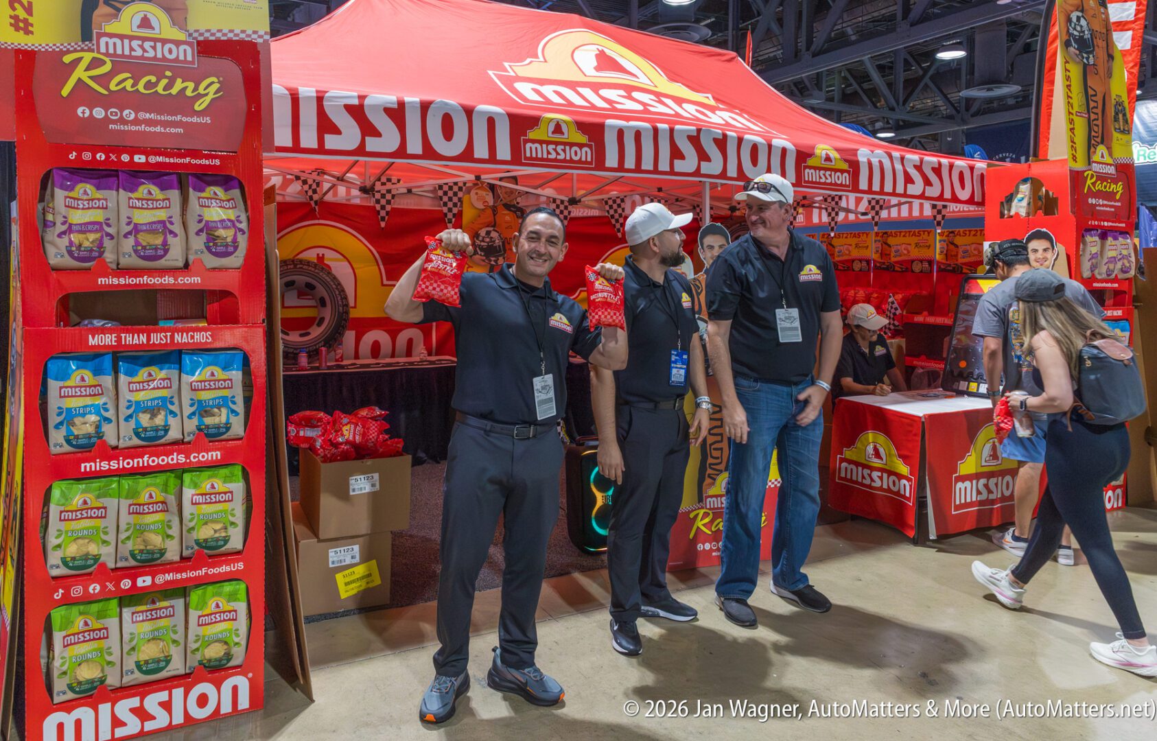 Three people stand and pose in front of a Mission Foods booth at an event, with product displays and a red Mission-branded tent in the background.