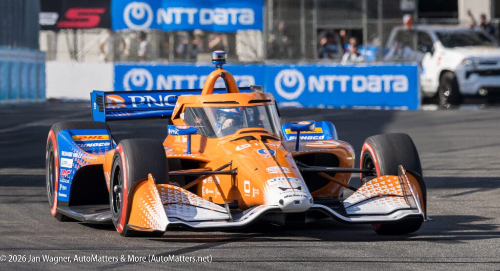 An orange and blue IndyCar races on a track during a motorsport event, with NTT DATA banners visible in the background.