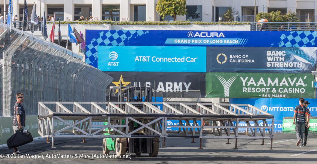 A forklift moves a section of metal bleachers on a race track, with workers nearby and sponsor banners displayed on the barriers in the background.