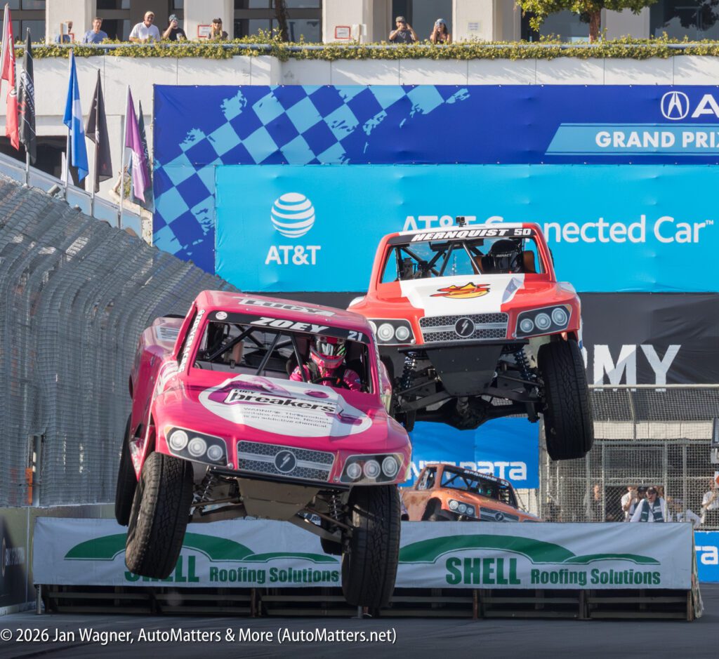 Two off-road trucks with sponsor logos jump side by side over a ramp during a stadium truck race, with crowd and banners in the background.