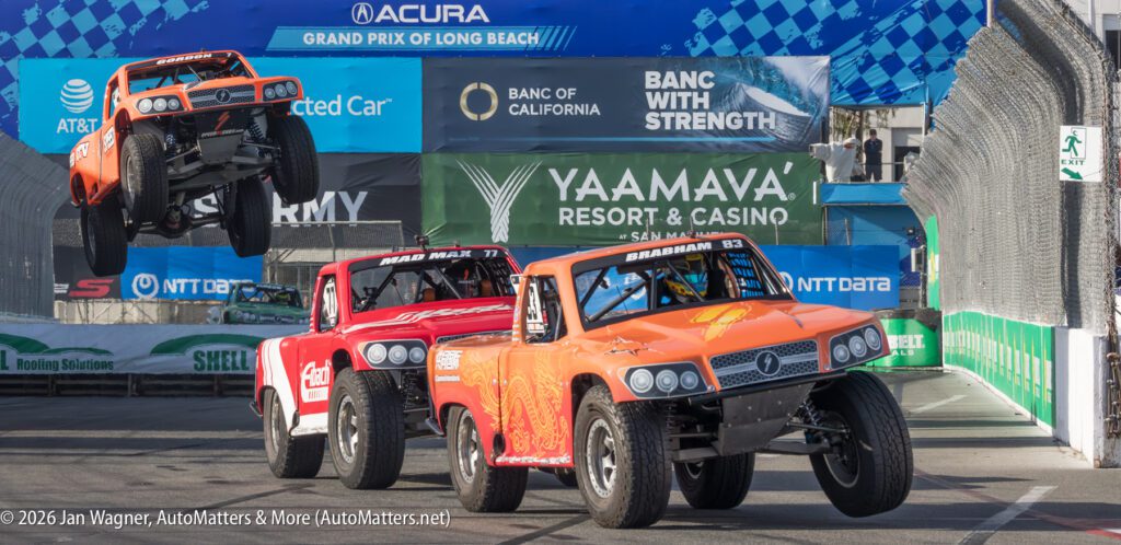 Three off-road racing trucks compete on a city street circuit, with one truck airborne over a ramp during the Acura Grand Prix of Long Beach event.