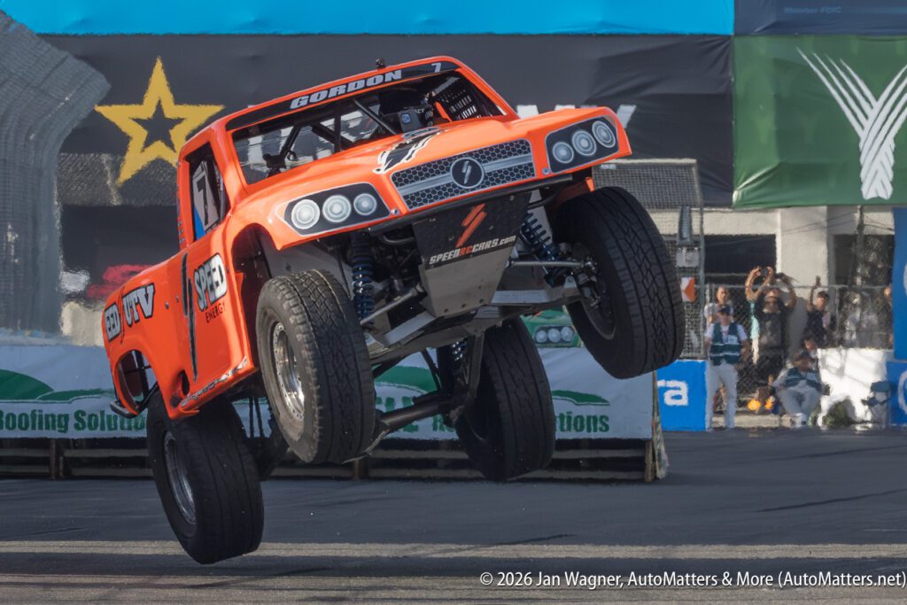 An orange off-road race truck lifts its front wheels off the ground while driving on a paved track during a motorsport event.
