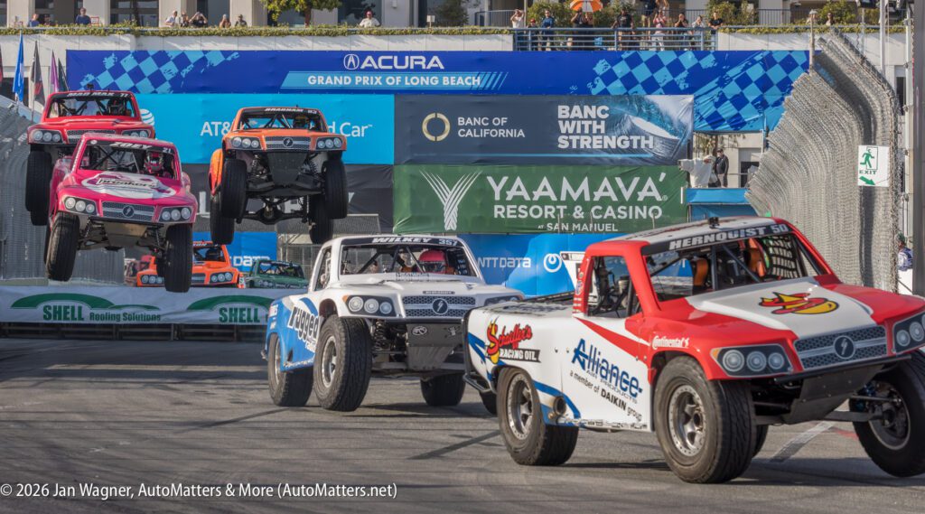 Several off-road trucks race on a city street circuit, with two trucks airborne over a ramp during the Grand Prix of Long Beach event.