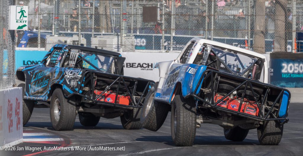 Two off-road racing trucks, tilted on two wheels, take a sharp turn on a street track during a motorsport event.