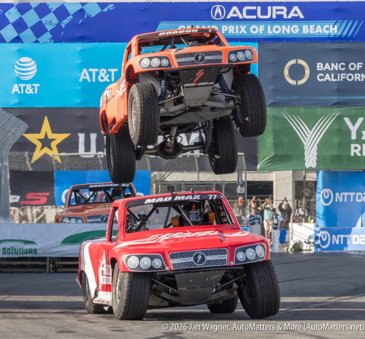 Two off-road trucks race on a street circuit; one truck is airborne above the other, both with wheels off the ground, in front of sponsor banners.