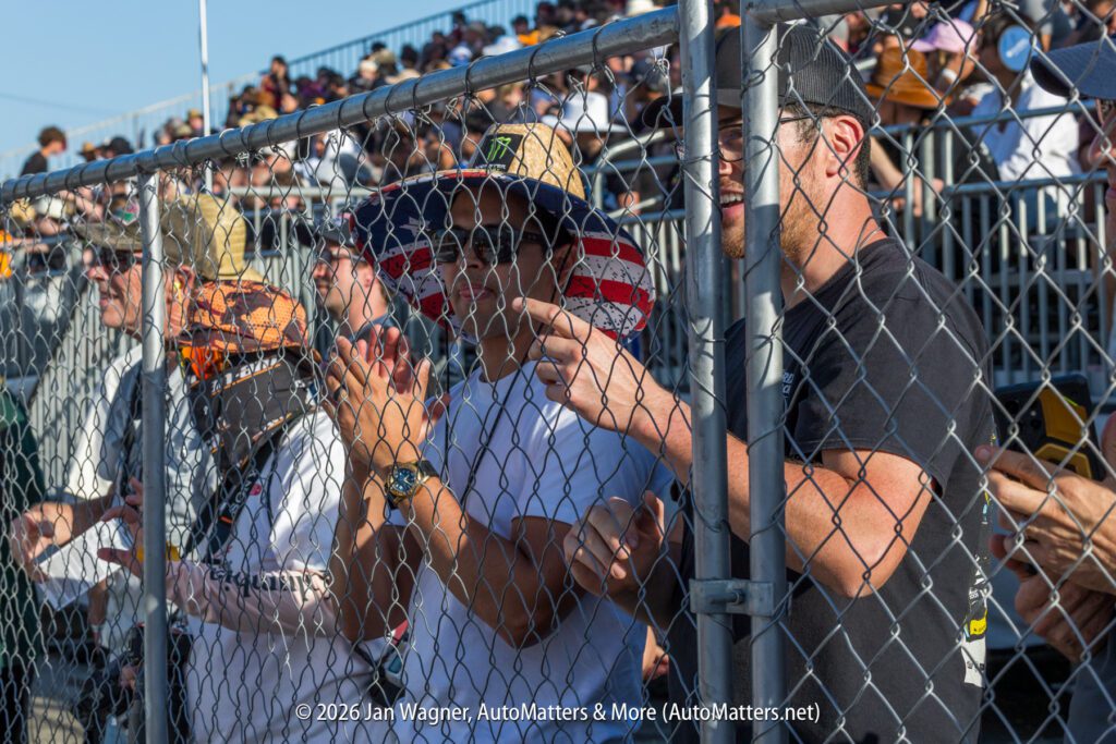 People stand behind a chain-link fence clapping and watching an outdoor event; one person wears a hat with an American flag pattern. Bleachers with seated spectators are visible in the background.