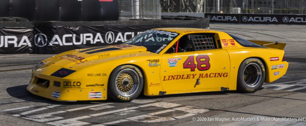Yellow Lindley Racing car number 48 crosses a racetrack finish line, surrounded by sponsor logos and a barrier with Acura branding.