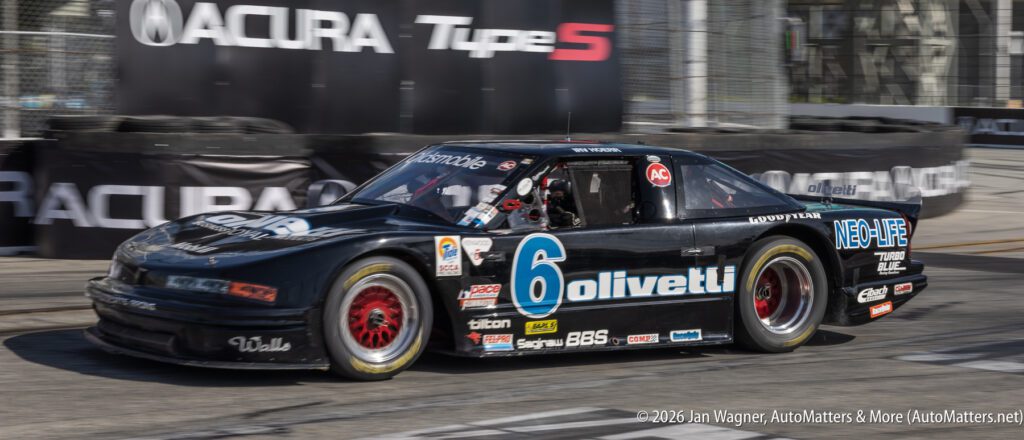 A black race car with the number 6 and various sponsor logos speeds on a racetrack, passing Acura signage in the background.