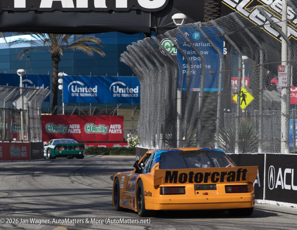 An orange Motorcraft race car drives around a track corner near a fenced barrier, with a green car and various sponsor banners visible in the background.