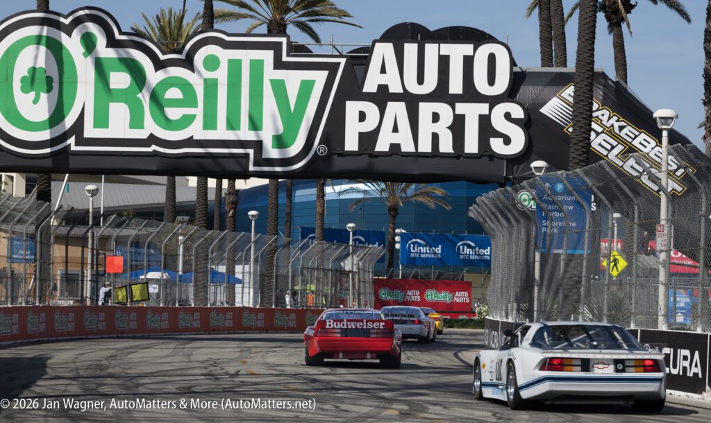 Three vintage race cars drive on a city street circuit under an O'Reilly Auto Parts sign, surrounded by fencing and palm trees.