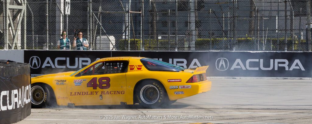 A yellow Lindley Racing car number 48 drifts and emits smoke on a racetrack near an Acura-branded barrier, with two people standing in the background.