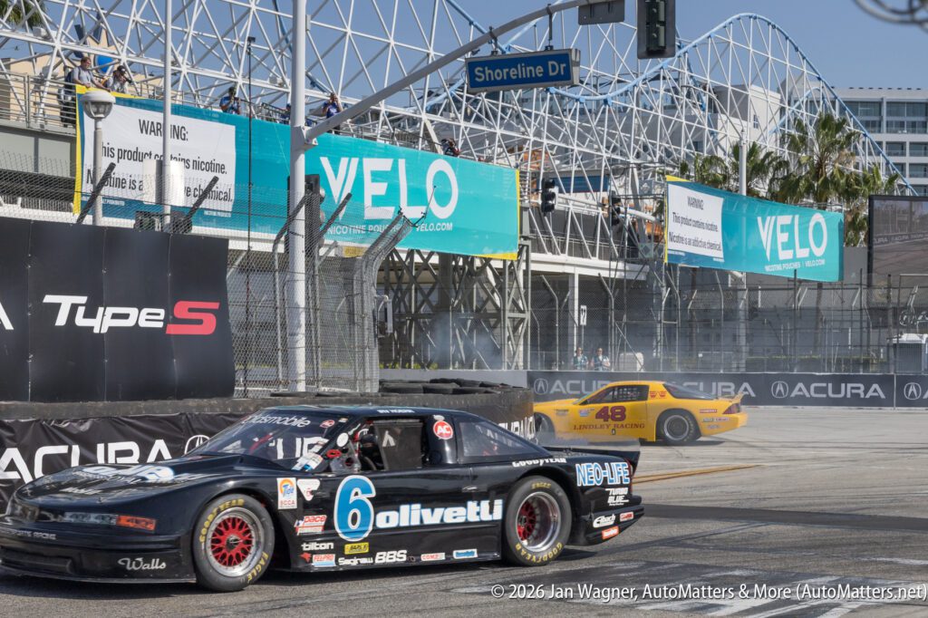 Black race car number 6 on a city racetrack passes a yellow car number 48, which is trailing smoke near a corner, with grandstands and advertising banners in the background.