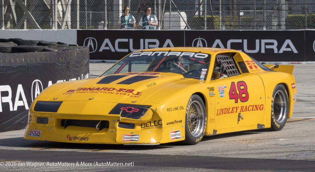 A yellow Chevrolet Camaro race car, number 48, with various sponsor logos, parked on a race track near a barrier, with Acura branding in the background.