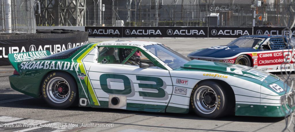 Green and white race car number 03 is parked on a race track next to a blue and red race car; both have prominent sponsor logos and are surrounded by fences and advertising banners.