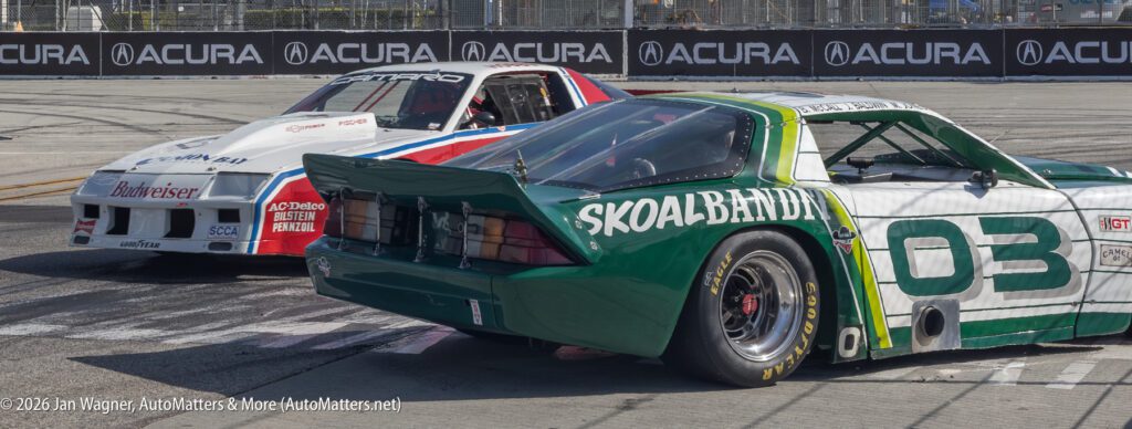 Two vintage race cars, one white with Budweiser branding and one green with Skoal Bandit branding, are on a racetrack near a barrier with an Acura sign.