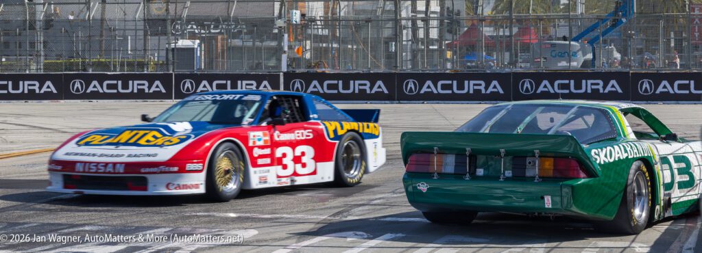 Two vintage race cars, one red and white #33 Nissan and one green #35 car, compete on a street circuit with Acura advertising banners in the background.