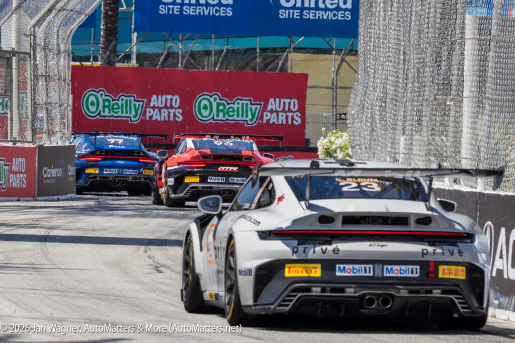A group of race cars drives closely together on a city street circuit, surrounded by safety barriers and O'Reilly Auto Parts banners.