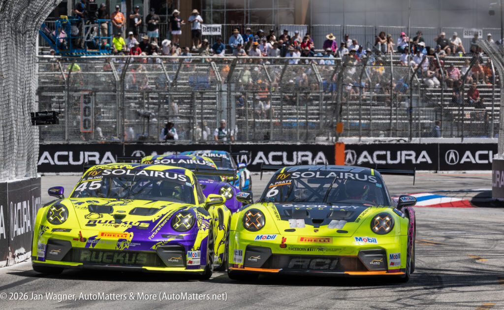 Two brightly colored race cars, number 45 and 17, compete side by side on a city track, with spectators watching from the stands behind a chain-link fence.