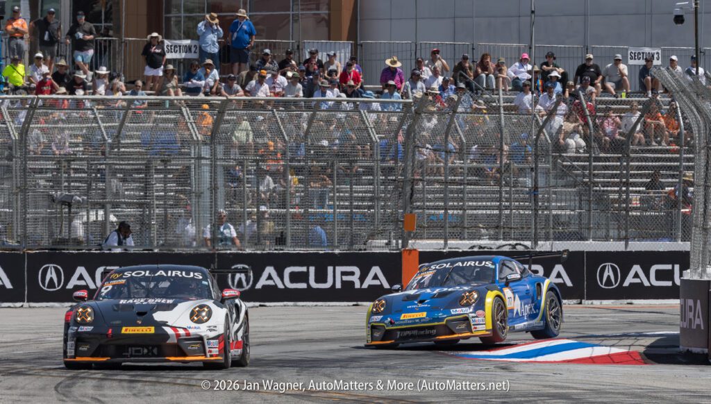 Two race cars compete on a track in front of a crowd behind a fence, with visible Acura and SOLAIRUS branding.