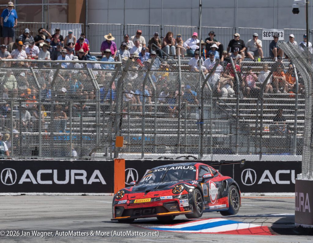 A black and red race car drives on a city circuit with barriers, track-side advertisements, and a grandstand filled with spectators in the background.