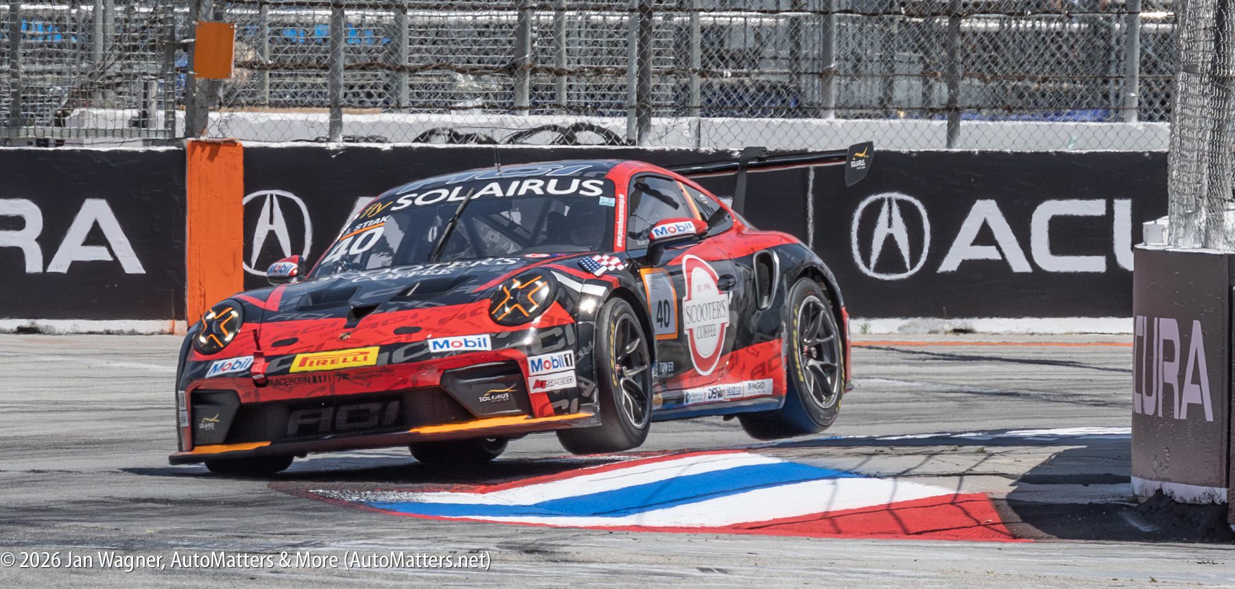 A race car lifts its front wheels off the ground while driving over a curb on a city track, with tire barriers and sponsor logos visible in the background.