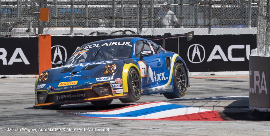 A blue Apex race car, number 17, drives over a curb on a racetrack during a race, with tire marks and safety fencing in the background.