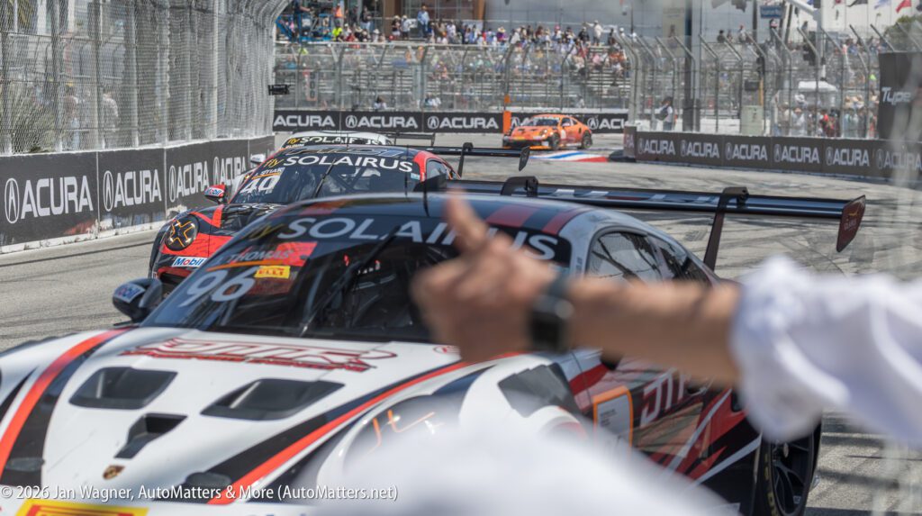 Race cars speed around a track near a fenced grandstand, with a person’s hand giving a thumbs-up in the foreground.