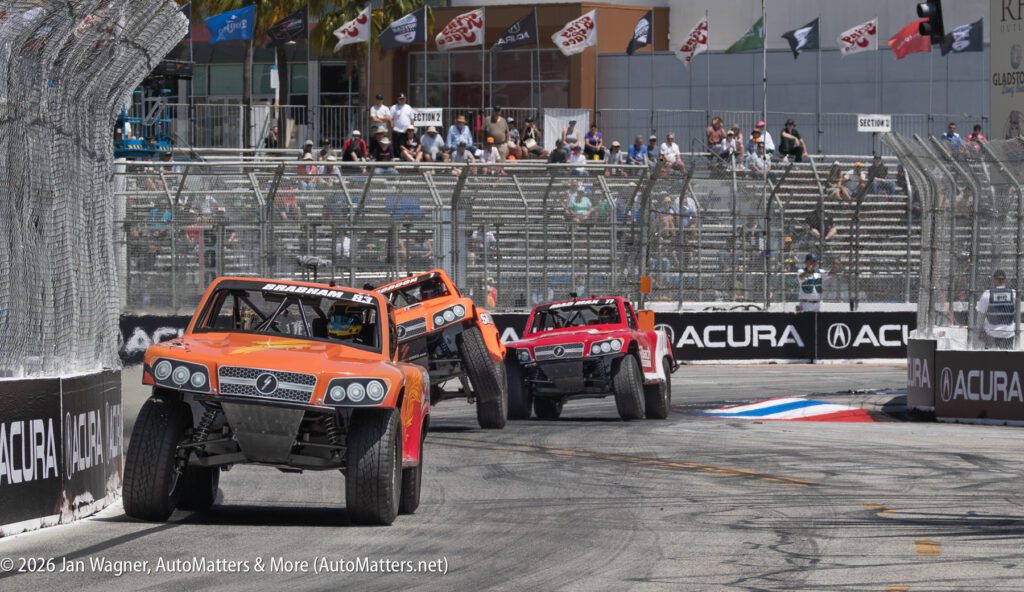 Several off-road trucks race closely on a city street circuit, passing barriers and taking a sharp turn, with spectators watching from behind fences.
