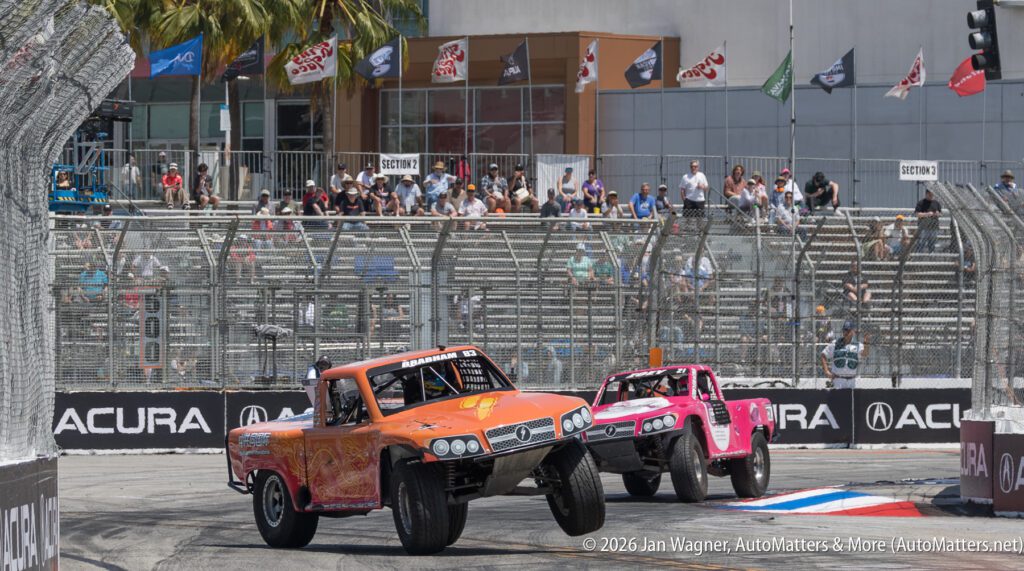 Two off-road trucks, one orange and one pink, race on a city street circuit, both with front wheels lifted, as spectators watch from bleachers behind a safety fence.