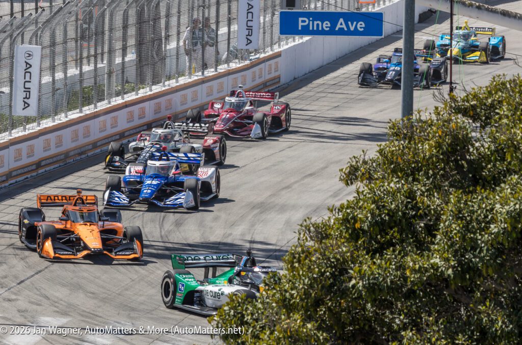 Several IndyCar race cars navigate a corner on a city street circuit with fencing, signage, and a "Pine Ave" street sign visible.