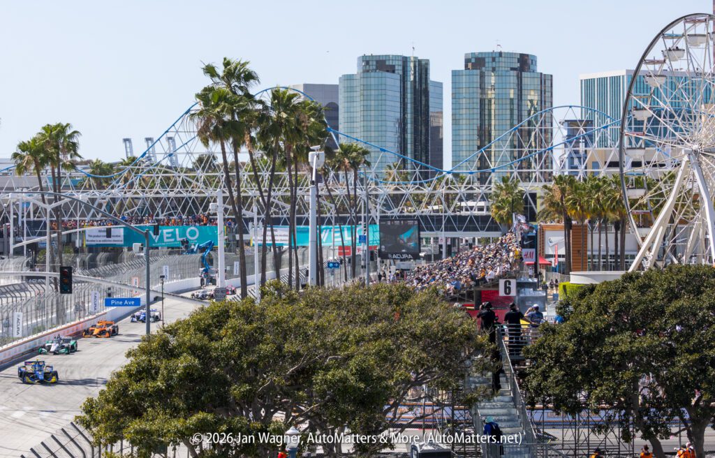 View of a street race event with race cars on track, spectators in stands, palm trees, and tall buildings in the background. Copyright information visible at bottom.