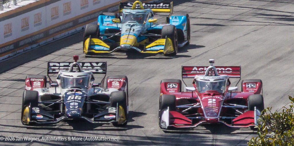 Three IndyCar race cars compete on a city street circuit, with two cars side by side in front and a third yellow-blue car closely behind.