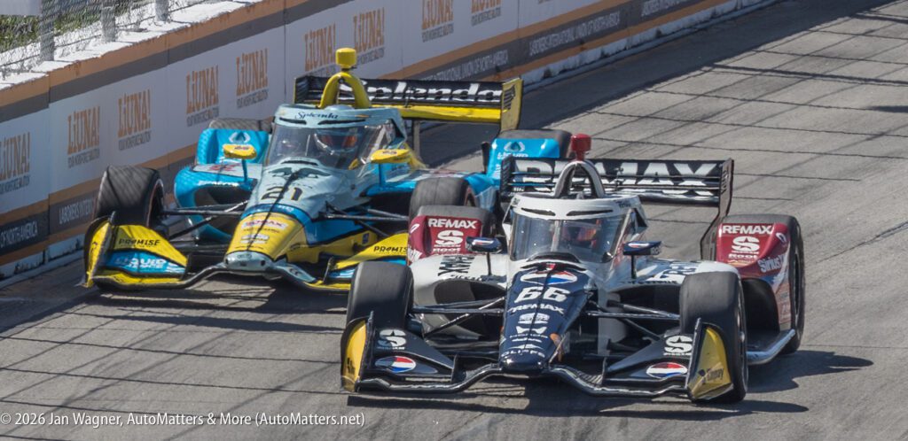 Two IndyCar race cars compete closely on a street circuit, with the black car #66 ahead of the blue and yellow car #21, both taking a tight corner.
