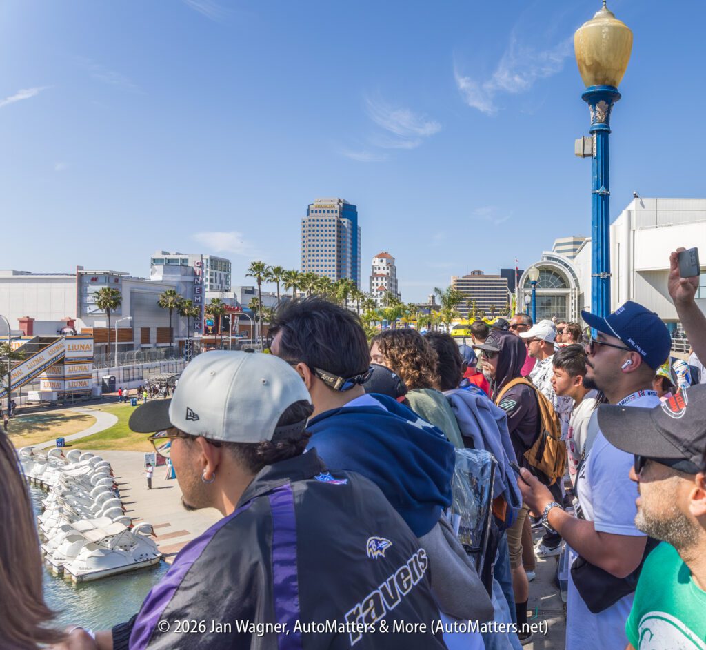 A crowd of people stands along a waterfront railing, looking out at water features and city buildings under a sunny sky.
