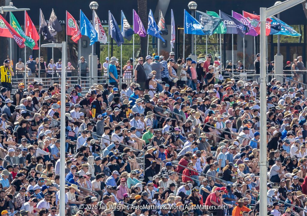 A large crowd of spectators sits in grandstand seats at an outdoor event, with colorful flags displayed above them.