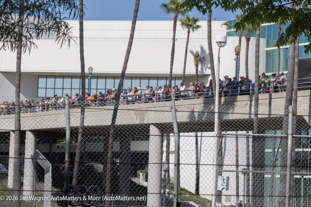 A large crowd stands on an elevated walkway outside a modern building, with palm trees and a chain-link fence in the foreground.