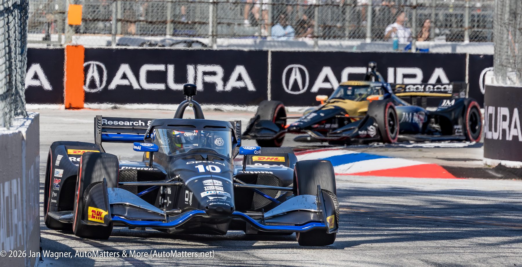 Two IndyCar race cars navigate a turn on a city street circuit, with track barriers and Acura branding visible in the background.