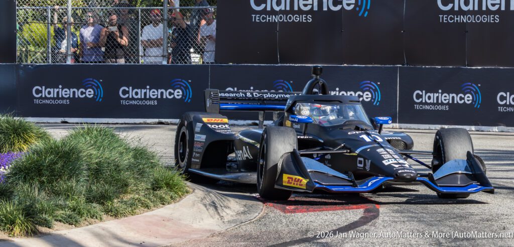 A black IndyCar with number 77 takes a corner on a street circuit during a race, passing by spectators and advertising banners for Clarience Technologies.