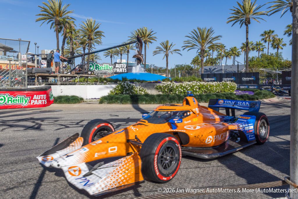 An orange and blue IndyCar speeds past O'Reilly Auto Parts signage on a street circuit lined with palm trees under a clear sky.