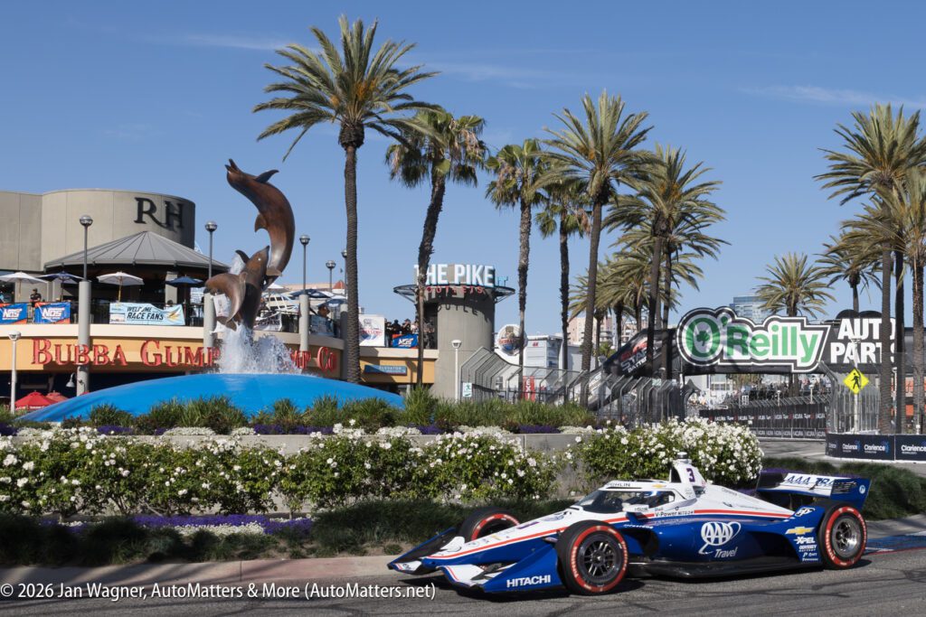 A blue and white IndyCar race car drives past palm trees, a dolphin fountain, and businesses including Bubba Gump Shrimp Co. and O’Reilly Auto Parts.