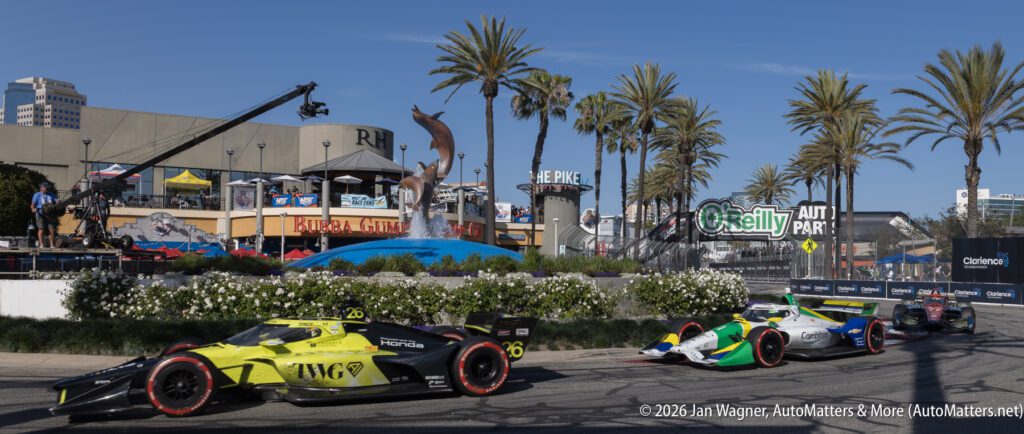 Three open-wheel race cars speed past palm trees and a water fountain during a street circuit race, with restaurants and an O'Reilly Auto Parts sign in the background.