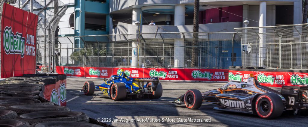 Two open-wheel race cars navigate a tight street circuit corner, surrounded by barriers with O'Reilly Auto Parts advertisements.