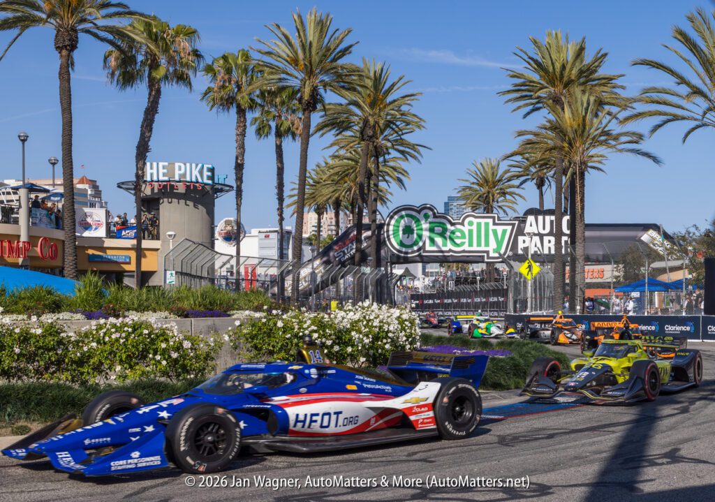 Indy cars race on a palm tree-lined street circuit near O'Reilly Auto Parts signage at The Pike in Long Beach, California, under clear blue skies.