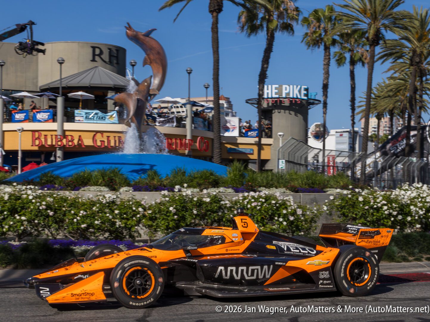 An orange and black Arrow McLaren IndyCar races past a dolphin fountain and the Bubba Gump Shrimp Co. restaurant at The Pike Outlets in Long Beach, California.