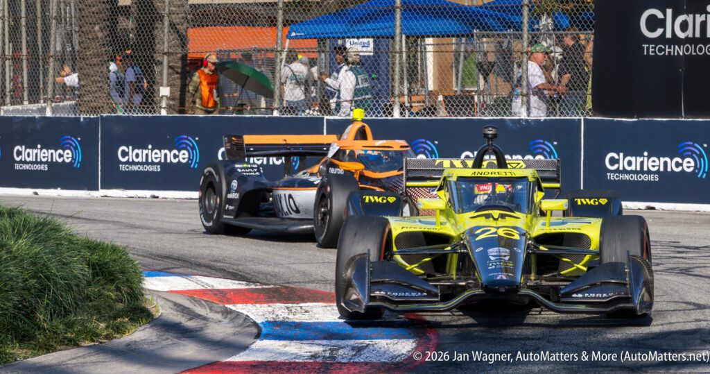 Two IndyCar race cars drive through a corner on a street circuit, with spectators and fencing in the background.