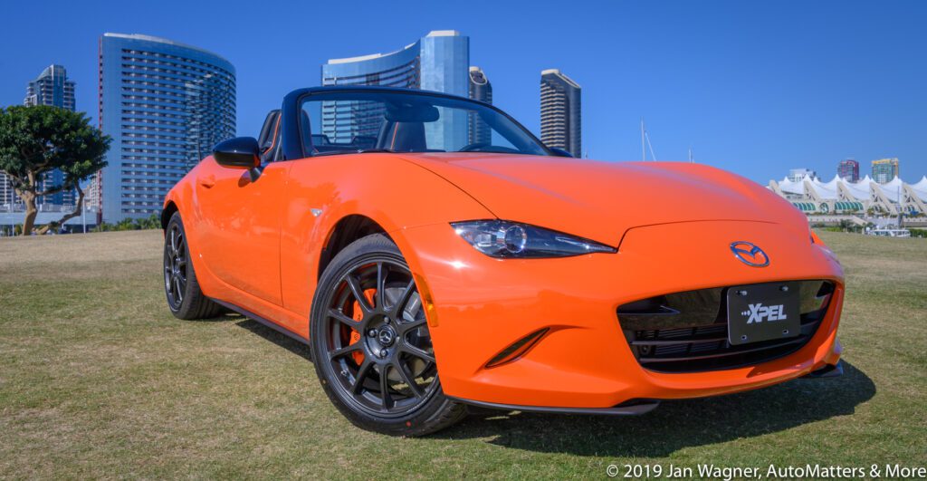 Bright orange Mazda MX-5 Miata convertible parked on grass with modern city buildings in the background under a clear blue sky.