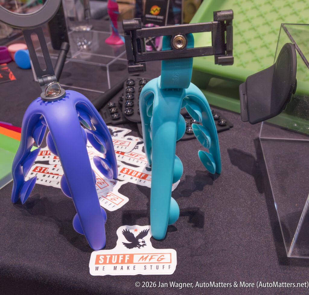 Two tripod phone holders, one blue and one teal, are displayed on a table with stickers that read “STUFF MFG WE MAKE STUFF.” Various tech accessories are visible in the background.