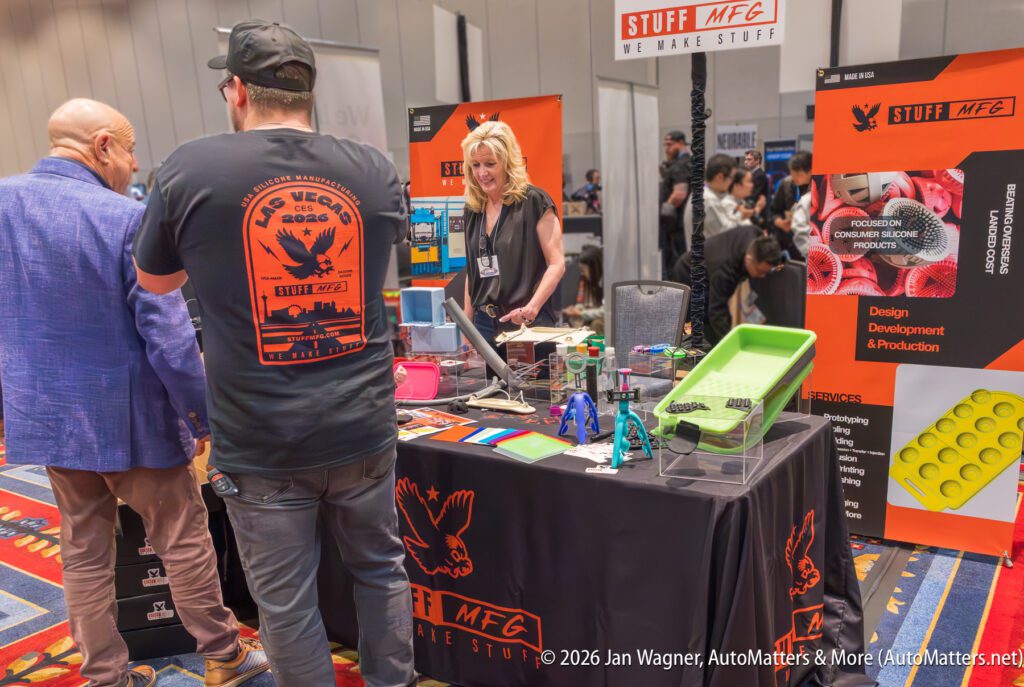 People interact at a trade show booth featuring product displays, informational banners, and 3D-printed items. A woman stands behind the table, engaging with attendees.