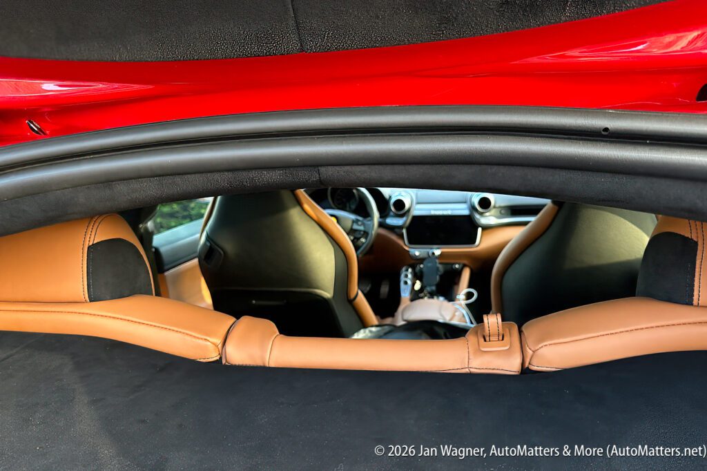 View through the open rear hatch of a red sports car, showing tan leather interior, front seats, and dashboard controls.